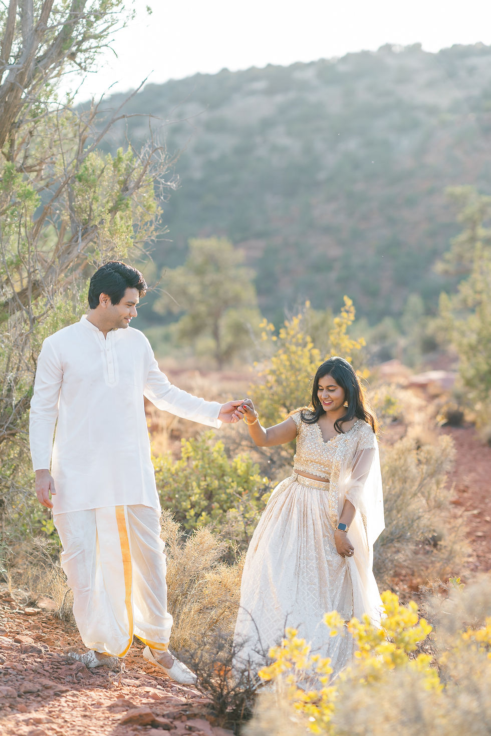 A couple in traditional attire holds hands on a rocky path, surrounded by greenery and hills. Bright, sunny day with a joyful mood.