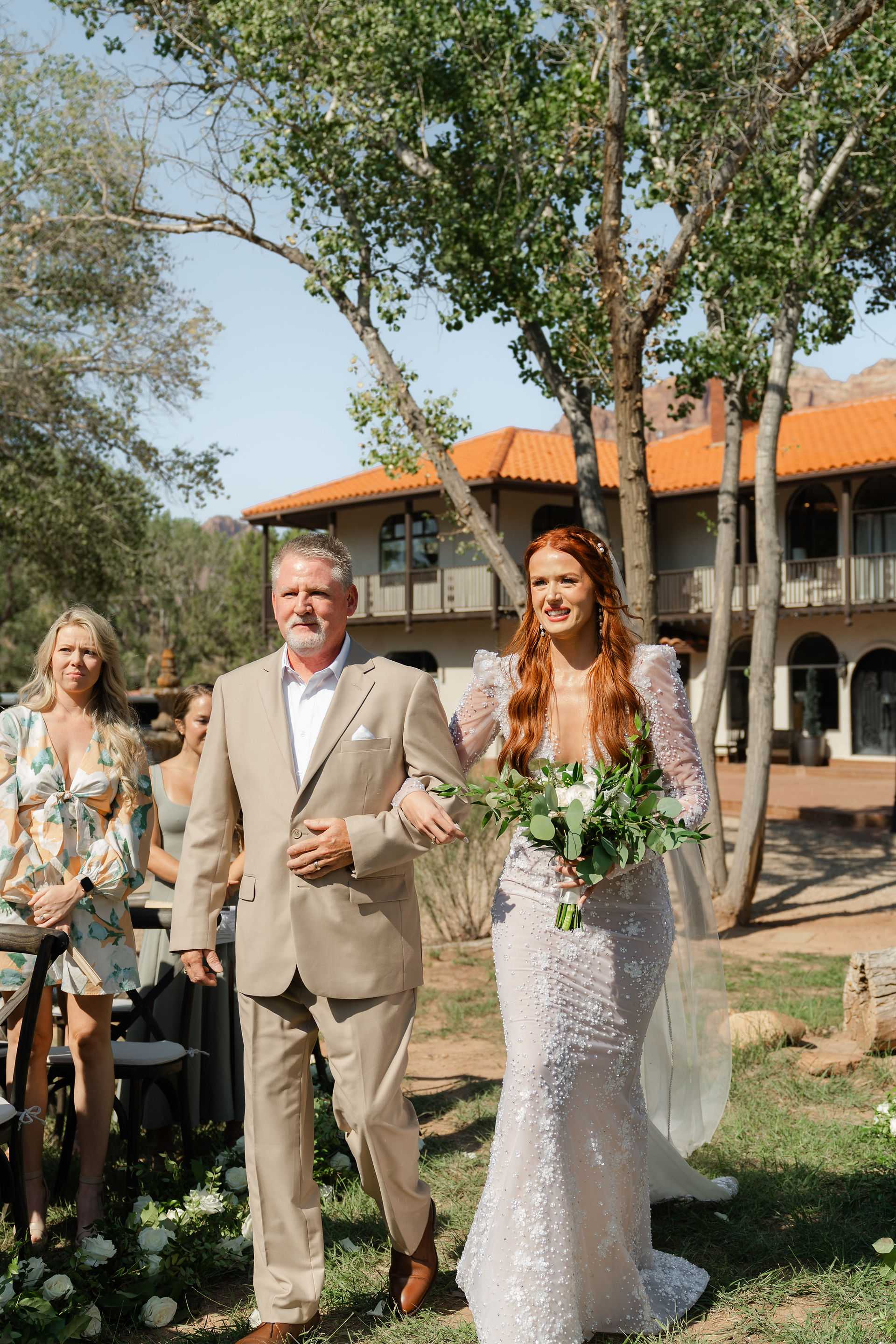 The bride, Laurel, beaming with joy as her father walks her down the sun-drenched outdoor aisle at her Zion National Park Wedding.