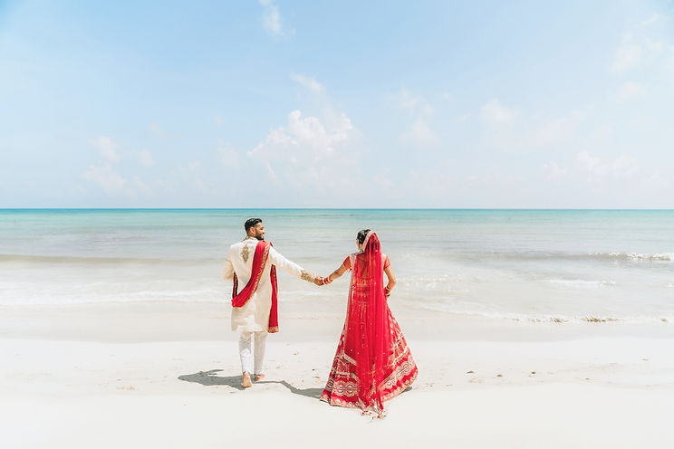 A South Asian groom in a traditional white and red sherwani and a bride in a red lehenga holding hands and walking along a white sand beach in Cancún, Mexico.