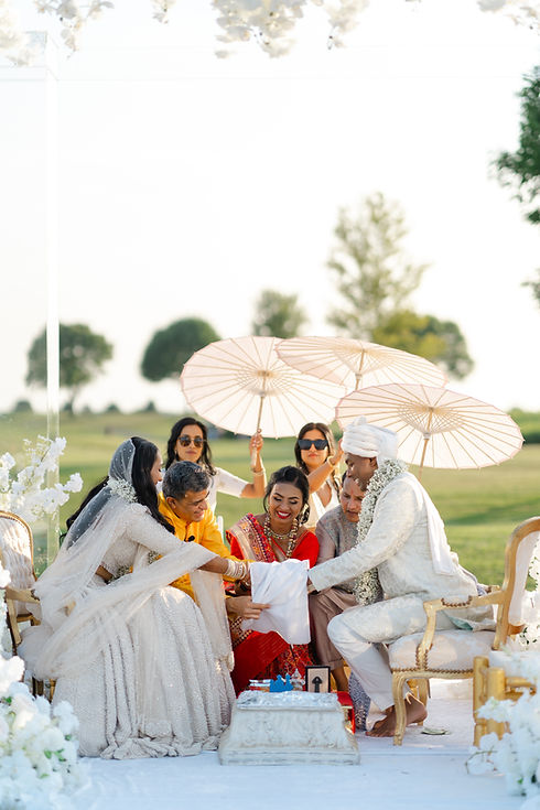 A South Asian groom and bride in traditional white and red wedding attire participating in a ceremony under a white parasol with guests in the background.