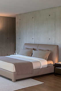 Chambre moderne avec lit beige, mur en béton, tapis blanc et grande fenêtre donnant sur un jardin.