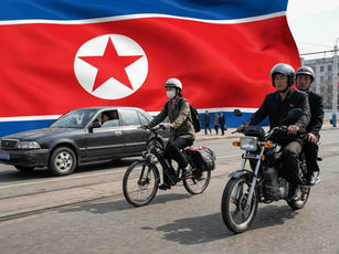 Riders on motorcycles and a bike in the street, passing a car. North Korean flag in the background. Busy urban setting.