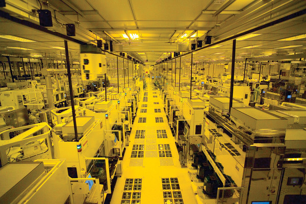 Yellow-lit semiconductor cleanroom with rows of automated machines and a central walkway, stretching into the distance.