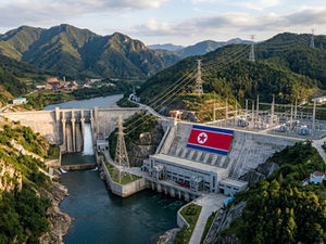 North Korean hydroelectric dam in a lush mountain valley, with power lines, water spillway, and a large DPRK flag on the wall.
