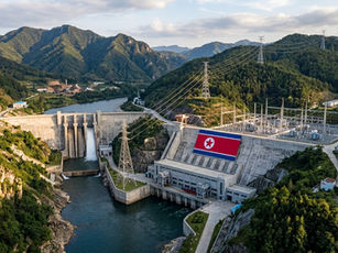 North Korean hydroelectric dam in a lush mountain valley, with power lines, water spillway, and a large DPRK flag on the wall.
