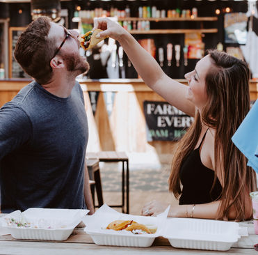 A woman playfully feeds a taco to a man at an outdoor eatery. They smile while sitting at a table with drinks and food in white plant-based containers.