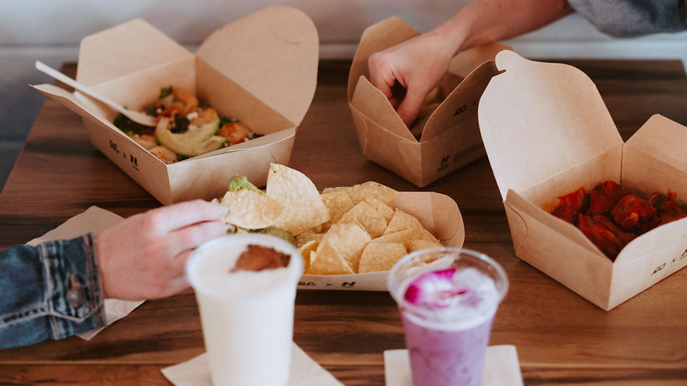 Hands reach for takeout, chips, and drinks on a wooden table. Compostable takeout boxes contain food; a purple drink with a flower in a compostable cold cup adds color. Casual setting.