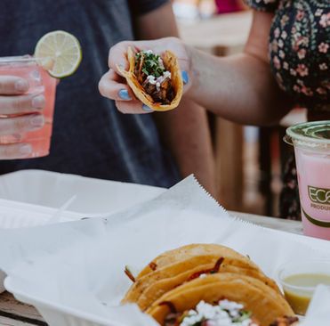 Two people holding tacos and drinks, one pink with lime, the other with a straw. Eco-branded cups and takeout boxes on a wooden table.