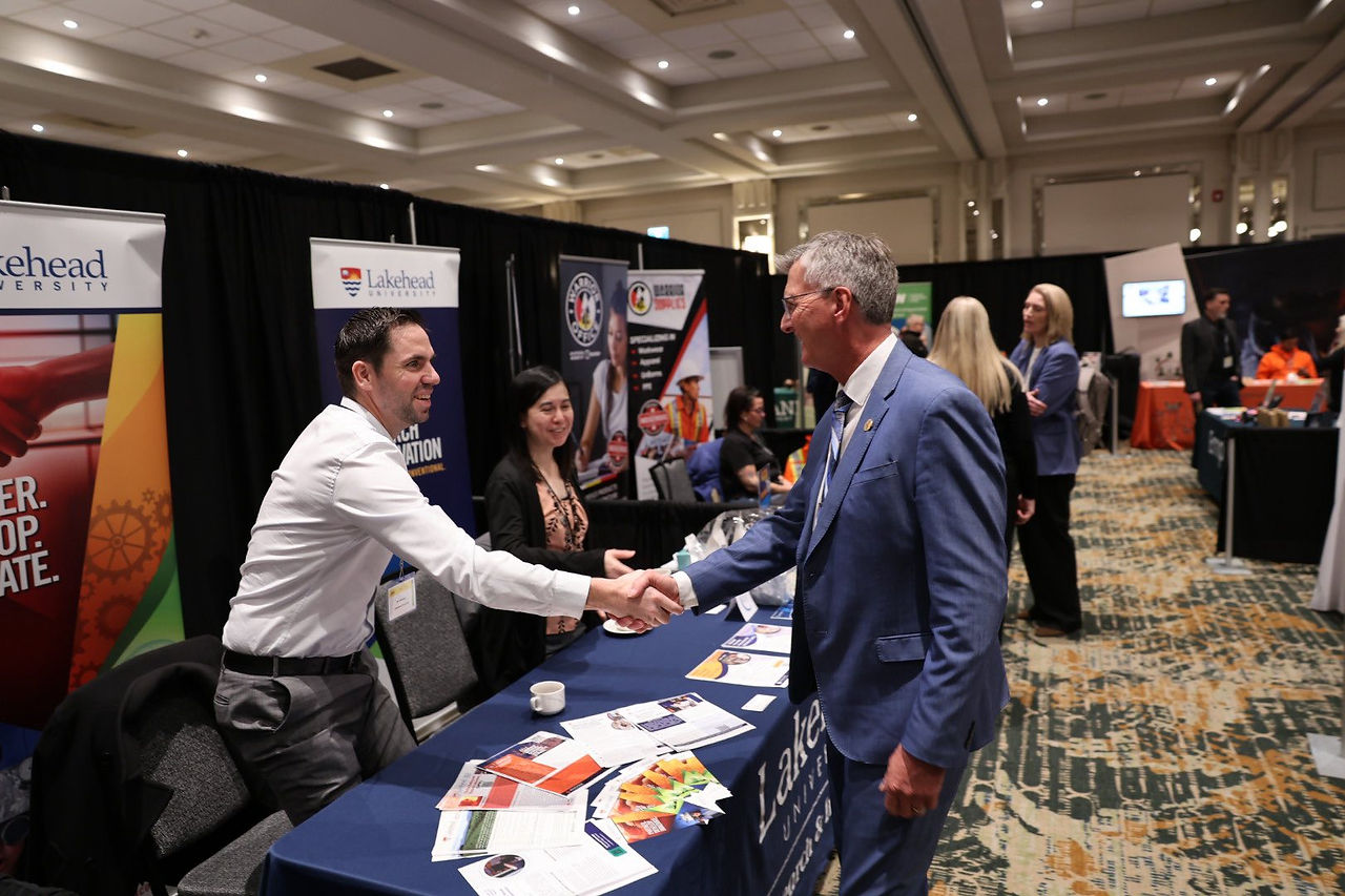 Businessmen shaking hands networking at a Lakehead University trade show booth.
