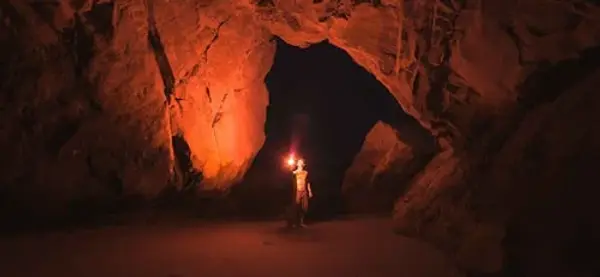 L'intérieur d'une vaste grotte rocheuse aux parois escarpées, éclairée par une lueur rouge orangé intense. Au centre, une silhouette humaine se tient debout dans l'obscurité, tenant une petite source de lumière vive qui crée un contraste saisissant avec l'entrée de la grotte en arrière-plan. Le sol semble sablonneux et plat, renforçant l'aspect monumental et mystérieux de la cavité.