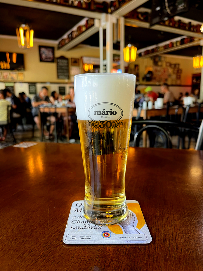 Copo de chopp claro com colarinho cremoso e brasão da Choperia do Mário, sobre mesa de madeira