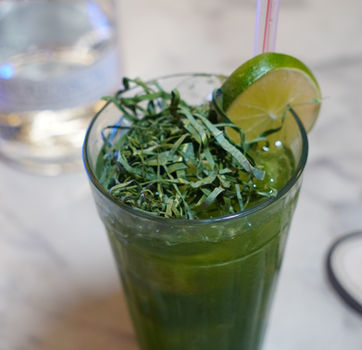 Copo de caipirinha verde com rapadura e couve, decorada com rodela de limão sobre balcão de mármore.