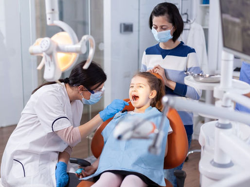 Dentist in blue gloves examines a child in a dental chair while her masked mother stands behind in a bright clinic.