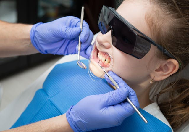 Child at dentist in blue bib and sunglasses gets teeth checked. Dentist's hands in purple gloves hold dental tools. Clinical setting.