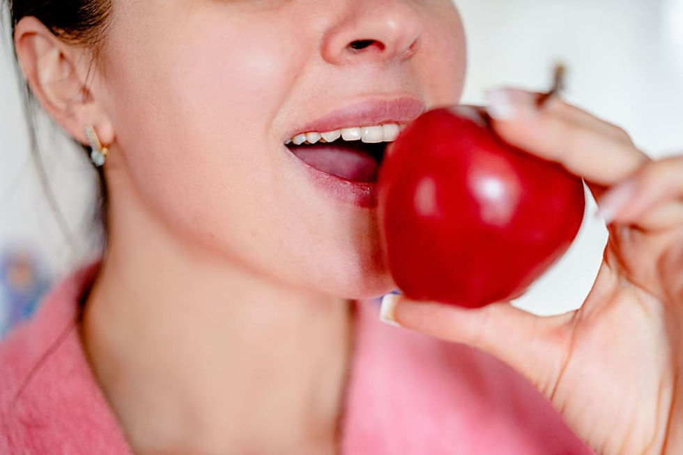 A person in a pink robe holds a shiny red apple close to their open mouth, ready to take a bite. The background is softly blurred.
