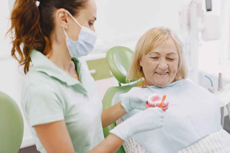 A dentist shows a senior woman a dental model in a clinic. The woman smiles, wearing a bib. The setting is bright with green dental chairs.