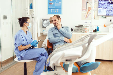 Dentist in blue scrubs consults a man in pain, holding his cheek, in a dental office. Posters and equipment are visible in the bright setting.