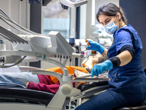 Dentist in blue scrubs and mask examines a patient in a dental chair. Bright lights and equipment visible, creating a clinical setting.