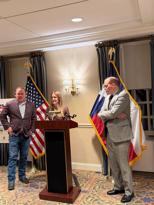Woman speaking at podium, two men beside her, flags in background