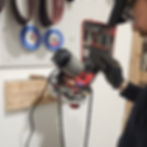 Person using a chainsaw sharpener on a mounted tool; sparks flying. Pegboard with tools and belts in the background.