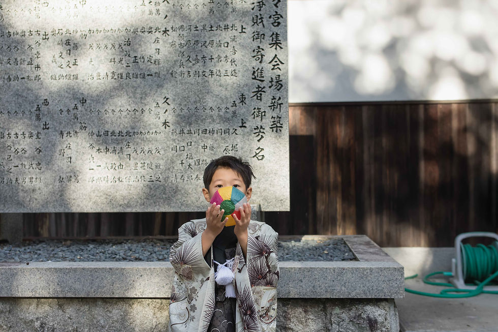神社の石碑の前で、和柄の着物を着た少年がカラフルな紙風船を顔の前に持ち、静かな雰囲気の中で立っている