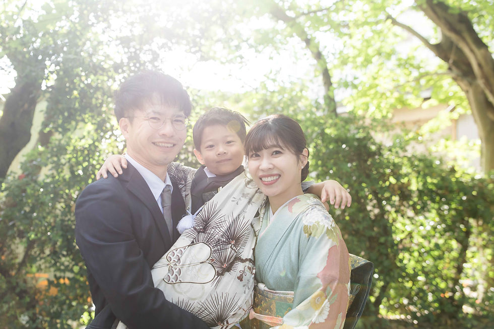 日差しの差す庭で、和装の両親に抱かれた子どもが笑顔の家族写真。緑の木々が背景に広がる。