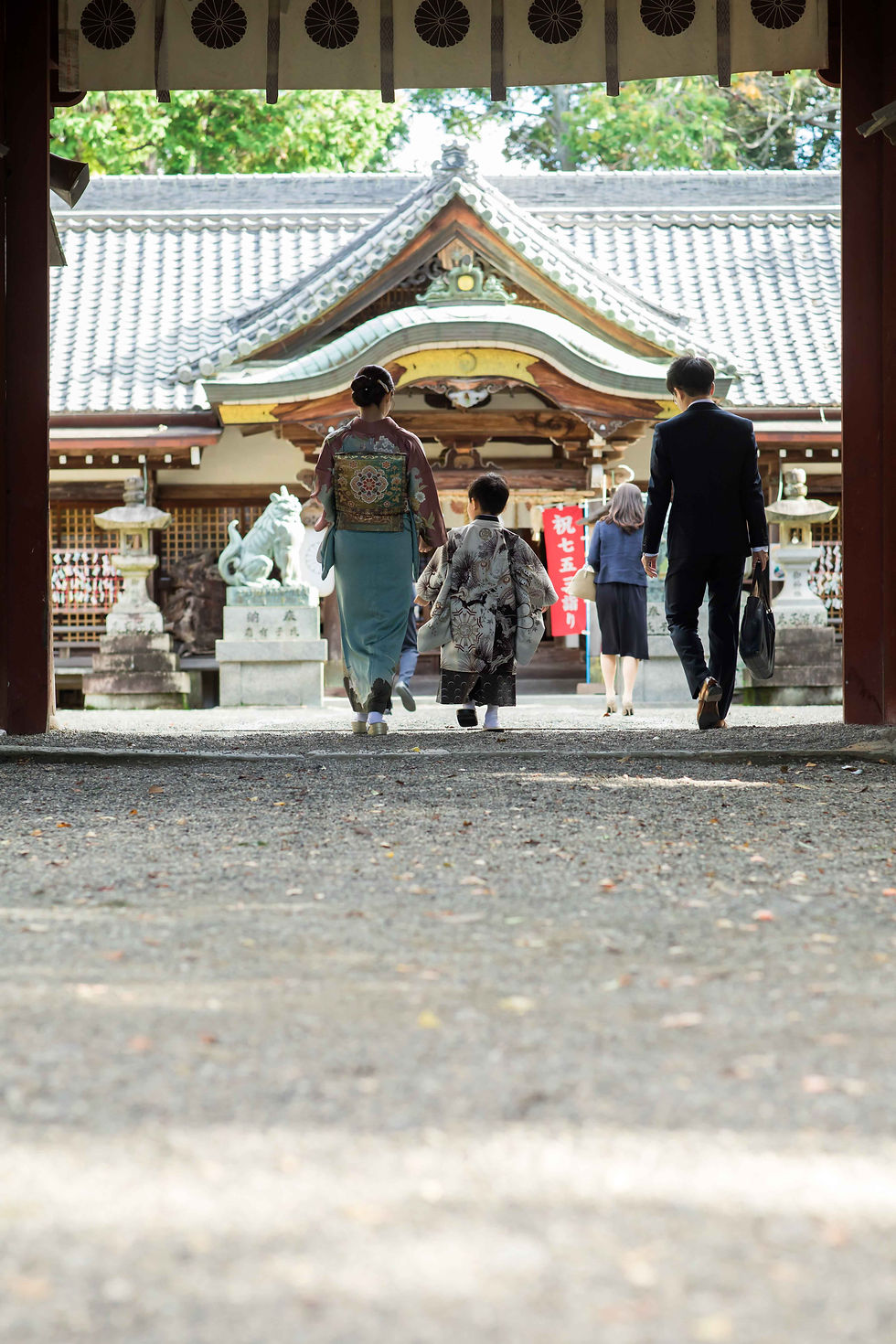 神社の参道を、着物の母子とスーツ姿の父が本殿へ向かう。紅白の「祝七五三詣り」札が見える、穏やかな晴れの日。