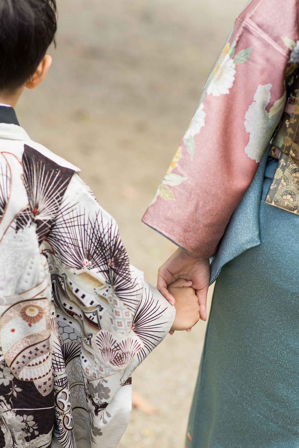 七五三の子どもが大人の手を握り、後ろ姿で並んで歩く。花柄や扇模様の着物が見え、穏やかな雰囲気。