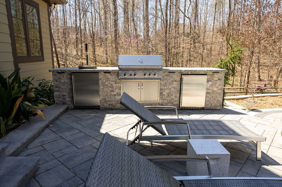Outdoor patio with stone grill station, stainless steel appliances, two lounge chairs, and a wooded backdrop. Peaceful and sunny.