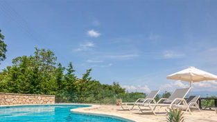 Бассейн с шезлонгами и зонтом под голубым небом среди деревьев.