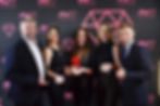 Five people smiling, holding awards against a black backdrop with pink "PM" logos and diamonds. Formal attire, celebrating success.