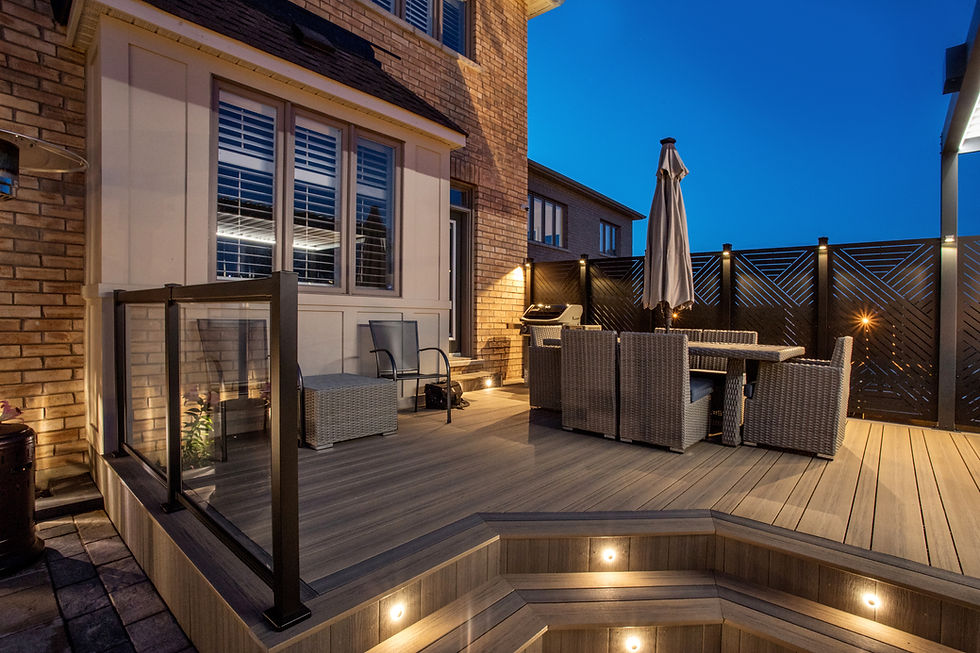 Evening view of a stylish composite deck with integrated LED step lighting, black aluminum and glass railings, a wicker dining set, privacy screen panels, and a barbecue setup against a brick home.