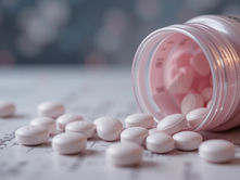A tipped-over prescription bottle with white pills spilled onto a surface, possibly a medical document or chart. The bottle label is partially visible, indicating medication. The background is blurred, emphasizing the pills and bottle, suggesting themes of medication use or healthcare.
