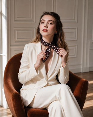 A woman in a white suit sits in a chair and adjusts a dark brown polka-dot bandana around her neck.
