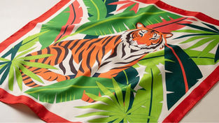 Close-up of a digitally printed silk-feel scarf featuring a bold tiger motif with lush green tropical leaves and a red border, elegantly draped to highlight its smooth fabric and vibrant colours.