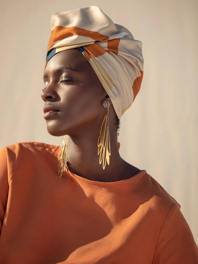 A woman with closed eyes wears a colorful headscarf and long gold earrings, dressed in an orange top.