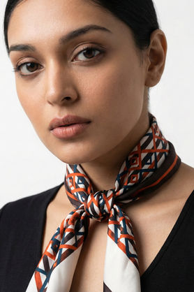 A woman wears a white and orange geometric print satin scarf tied in a neat neck knot over a black top.