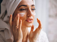 A person with a white towel wrapped around their head applies white cream to their face with both hands. They are wearing a light-colored robe, and the background is softly lit, suggesting a spa or skincare routine setting.