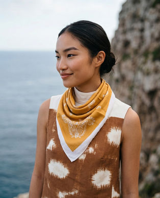 A woman smiles wearing a mustard paisley neck scarf against a blurred coastal cliff backdrop.