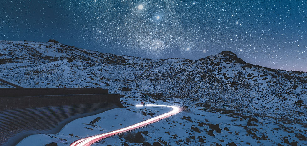 Snow covered mountains in the night time with star behind them. Road over the mountain is lit up by car headlights. 