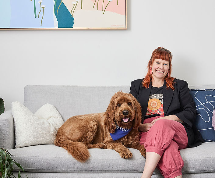 Woman with copper hair in black blazer and pink trouser sitting next to a big tan dog on a grey sofa in the waiting room