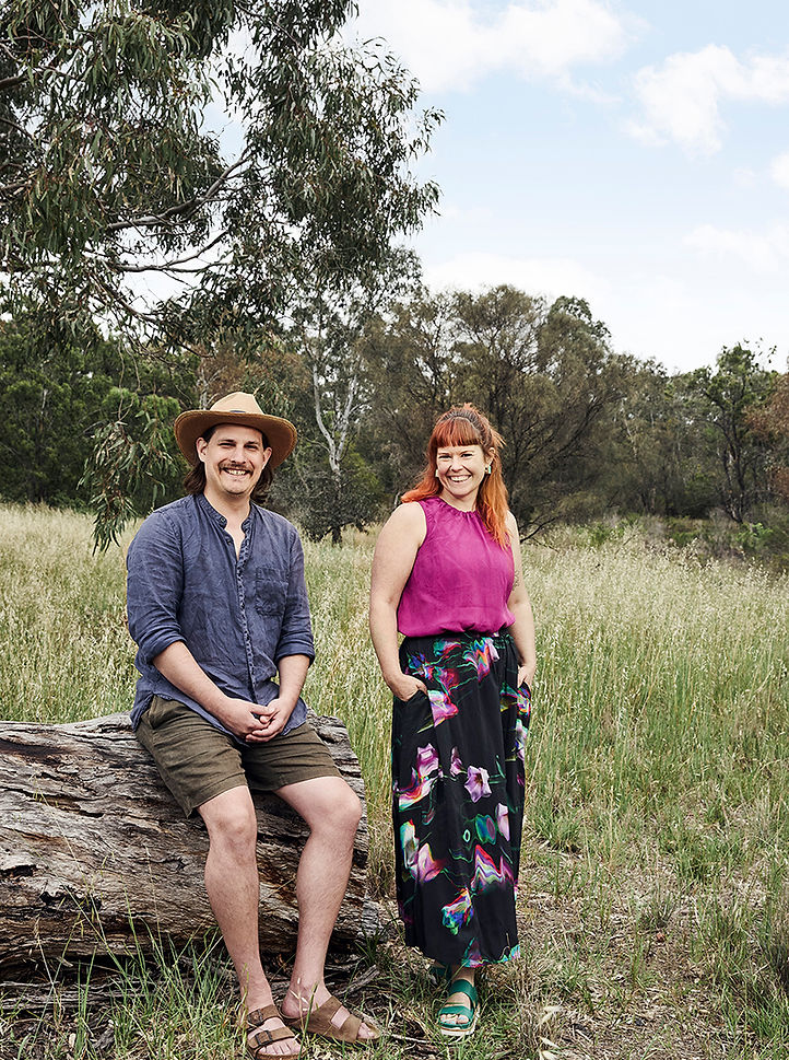 A main in a blue shirt and hat sitting on a log next to a woman with copper hair and pink singlet top and skirt in the bush