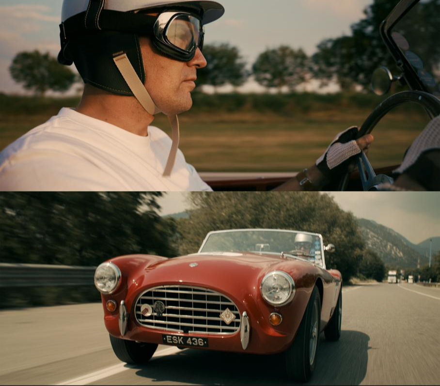 A man in a vintage racing helmet and goggles driving a red classic sports car