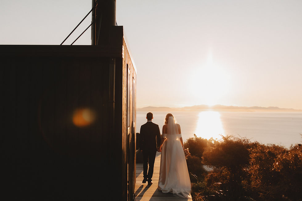 A wedding couple walk hand in hand at sunset by the sea, next to a dark building. The sky and water glow with warm light.