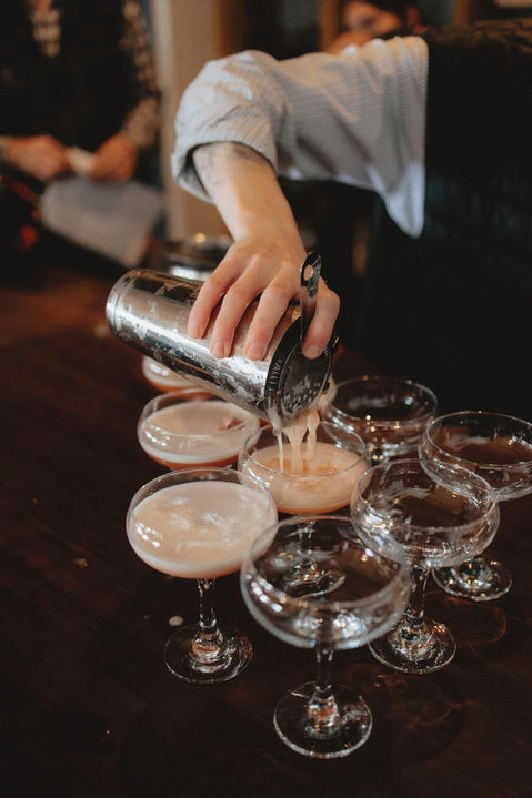 Hand pouring cocktail from shaker into several coupe glasses on a dark bar.