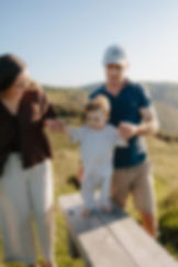 Baby taking assisted steps outdoors with parents on a wooden bench.