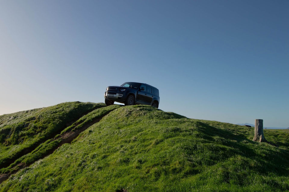 Dark Landrover Defender parked on a green grassy hill under blue sky.