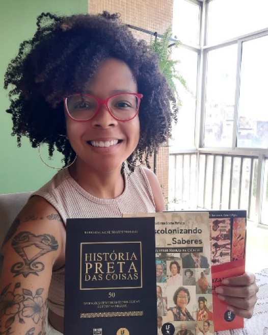 Bárbara carine, sorrindo para a câmera, segura os três livros de sua autoria