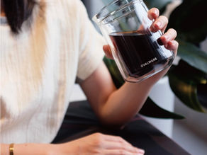 A person holds a glass mug of dark coffee labeled "Ovalware." They're sitting in a room with green plants, wearing a cream shirt and a gold bracelet.