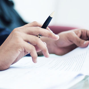 Hands holding a pen and a document with text, in a bright setting. The focus is on reviewing or editing the paperwork.